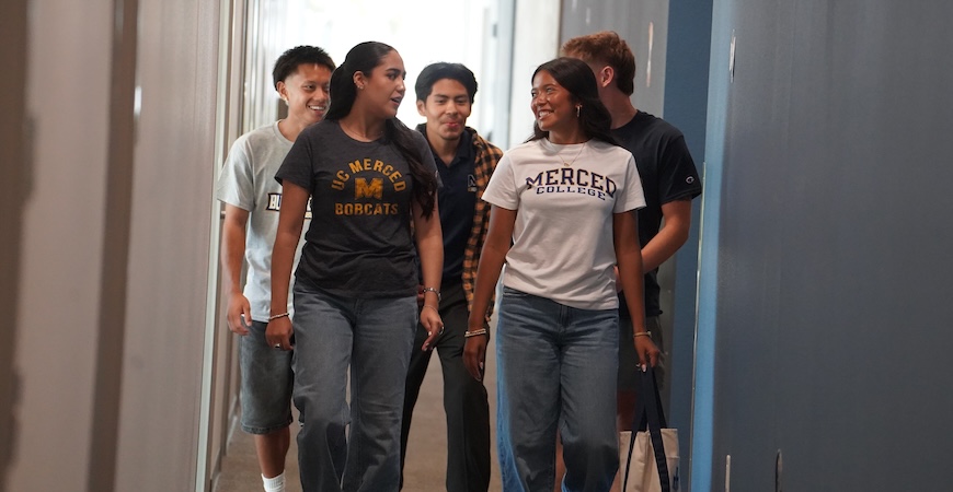 Students wearing shirts emblazoned with UC Merced and Merced College logos walk down a hallway.