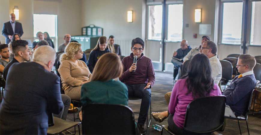 A male undergraduate student sits in a circle and speaks to people. 