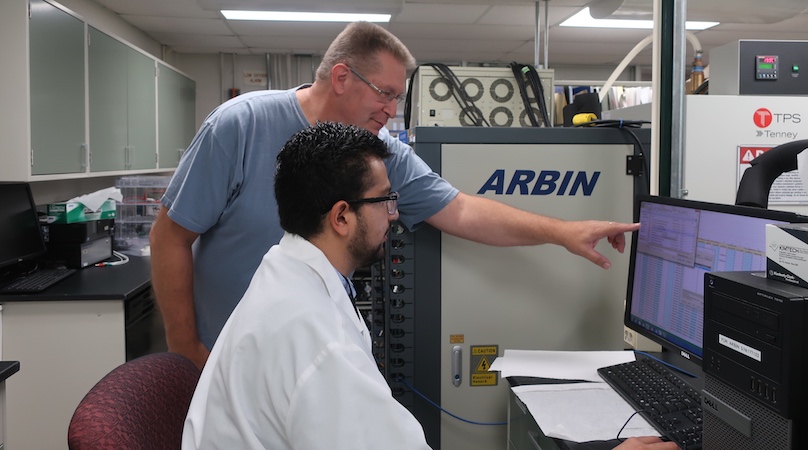 A student sitting at a computer with a NASA scientist standing behind him are working together in a laboratory.